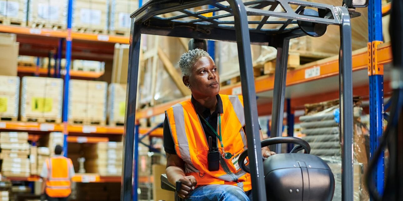A photograph of a Black female forklift driver with short grey hair wearing a hi-vis vest while driving a forklift truck between tall shelving units